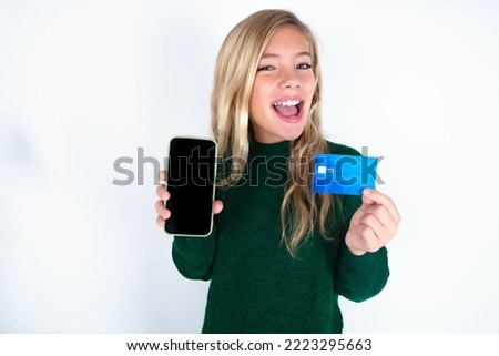caucasian teen girl wearing green knitted sweater over white background opened bank account, holding smartphone and credit card, smiling, recommend use online shopping application