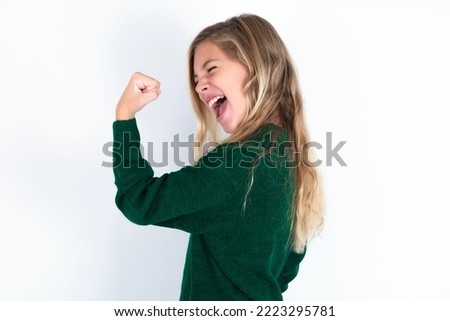 Portrait of funny caucasian teen girl wearing green knitted sweater over white background shout yeah raise fists hands celebrate victory game competition
