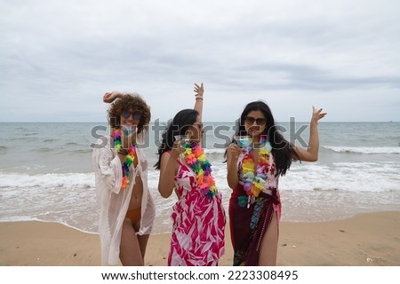 Attractive mature woman with two young, pretty, brunette South American women in bikinis, sunglasses and flower necklaces dancing with glasses of blue wine. Concept vacation, friends, summer, drinks.