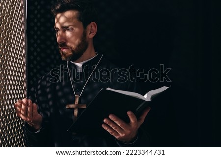 Catholic priest in cassock holding Bible and talking to parishioner in confessional Royalty-Free Stock Photo #2223344731