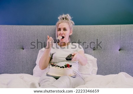 Bored, sad woman in home clothes eating cake and using the remote control to change the channel while watching television in bed at night. Overeating, Jamming of negative emotions. Selective focus.