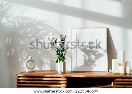 Modern minimalist Scandinavian style interior with white poster mockup, candles and flowers in vase on a wooden console under sunlight and home plants shadows on a white gray wall. Selective focus.