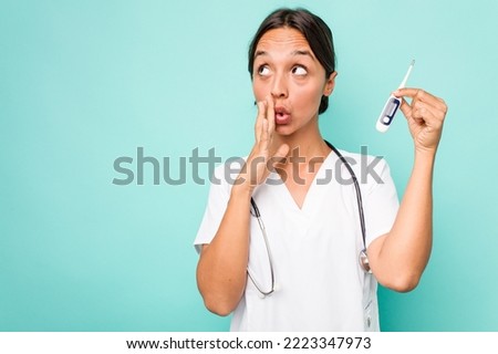 Young hispanic nurse woman holding a thermometer isolated on blue background is saying a secret hot braking news and looking aside