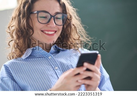 Smiling young professional business woman, happy businesswoman holding smartphone standing in office using mobile looking at cell phone, texting on cellphone, typing on cellular technology device.