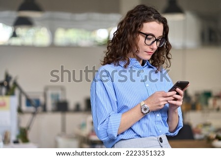 Busy young business woman manager holding smartphone working standing in office using mobile cell phone, checking corporate apps, browsing online on cellphone typing on cellular technology device. Royalty-Free Stock Photo #2223351323