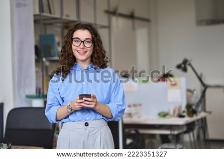 Smiling young business woman, happy businesswoman corporate leader holding cellular smartphone working standing in office using mobile cell phone working on cellphone looking at camera. Royalty-Free Stock Photo #2223351327