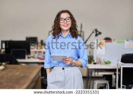 Young smart smiling professional business woman, happy confident 30s female company worker or sales manager holding digital tablet standing in modern office working, looking at camera, portrait.