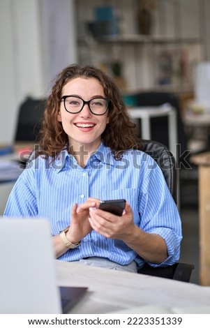 Young happy smiling professional business woman worker holding mobile phone using apps on cellphone working in office with smartphone browsing cellular technology, looking at camera, vertical.