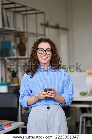 Smiling young business woman, happy businesswoman corporate leader holding cellular smartphone working standing in office using mobile cell phone working on cellphone looking at camera. Vertical Royalty-Free Stock Photo #2223351347