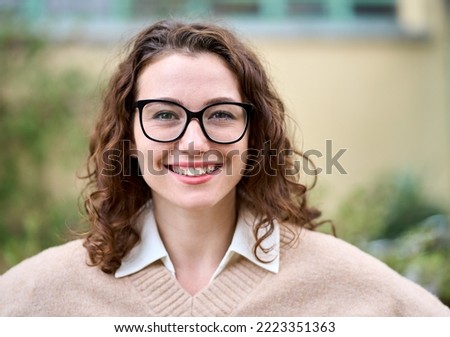 Young pretty smiling professional business woman, happy businesswoman, positive beautiful curly lady wearing glasses standing outdoor on street, looking at camera, front face headshot portrait. Royalty-Free Stock Photo #2223351363
