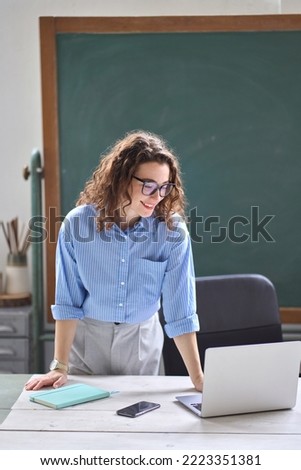 Young happy woman school professional teacher, online tutor standing at desk in front of chalkboard in classroom working on laptop computer teaching remote education virtual classes, vertical. Royalty-Free Stock Photo #2223351381