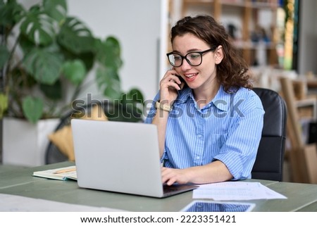 Young business woman professional employee, entrepreneur sitting at desk working online on laptop making call on cell phone taking to client regarding online website sales order or marketing work. Royalty-Free Stock Photo #2223351421