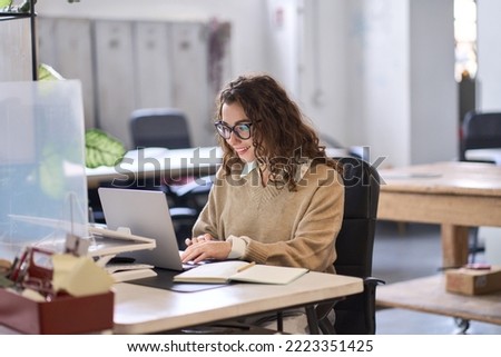 Young happy professional business woman employee sitting at desk working on laptop in modern corporate office interior. Smiling female worker using computer technology typing browsing web. Royalty-Free Stock Photo #2223351425