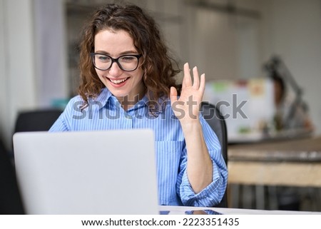 Young happy business woman using laptop waving hand greeting workers on remote virtual work conference meeting call in office. Hybrid video chat conversation corporate videoconference management. Royalty-Free Stock Photo #2223351435