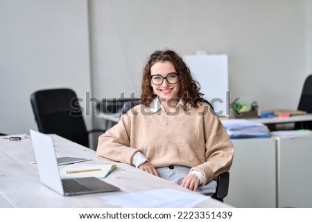 Young adult happy smiling professional business woman, female company office worker or businesswoman marketing sales manager sitting at work desk, looking at camera, posing for business portrait. Royalty-Free Stock Photo #2223351439
