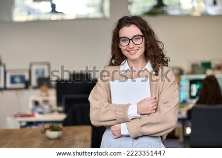 Young smiling professional business woman, happy businesswoman, female company worker intern or corporate manager holding laptop standing in modern office working, looking at camera. Portrait Royalty-Free Stock Photo #2223351447