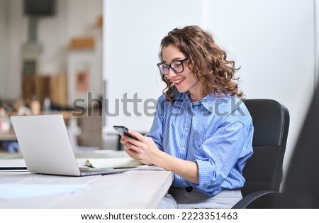 Young happy business woman, smiling beautiful professional businesswoman looking at smartphone using cellphone mobile cell technology working at home in office typing on cell phone sitting at desk. Royalty-Free Stock Photo #2223351463