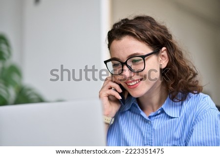 Young woman sales manager talking on cell phone using laptop working online on computer. Female professional entrepreneur making call consulting customer on mobile selling e services in office. Royalty-Free Stock Photo #2223351475