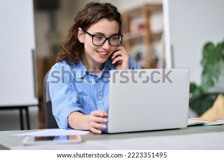Young woman sales manager talking on cell phone using laptop working online on computer. Female professional entrepreneur making call consulting client on mobile selling web services in office. Royalty-Free Stock Photo #2223351495