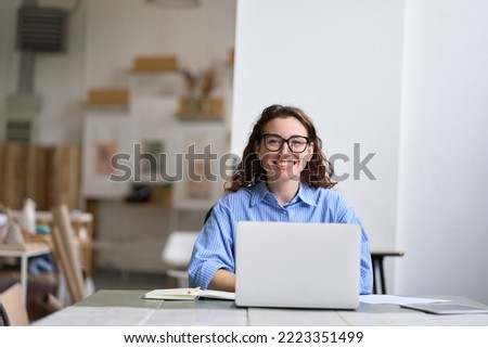 Young happy business woman company employee sitting at desk working on laptop. Smiling female professional designer or student using computer in corporate office looking at camera, portrait.