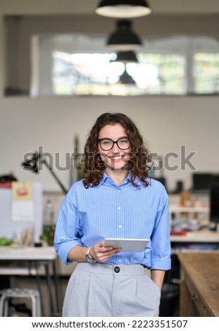 Young smart smiling professional business woman, happy confident 30s female company worker sales manager holding digital tablet standing in modern office working, looking at camera, vertical portrait. Royalty-Free Stock Photo #2223351507