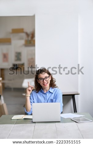 Young happy business woman sitting at desk working on laptop having idea or solution. Smiling female professional worker or student using computer in corporate office pointing finger up in coworking. Royalty-Free Stock Photo #2223351515