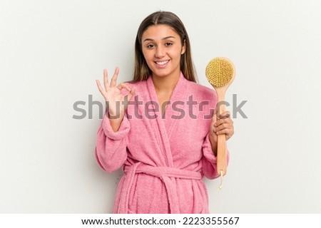 Young Indian woman holding a shower brush isolated on white background cheerful and confident showing ok gesture.