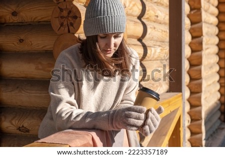 A brooding young woman stands on the terrace of a log house with a glass of coffee. Sunny autumn day. High quality photo