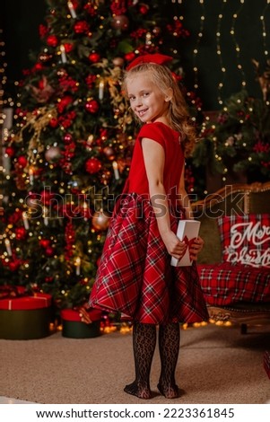 Pretty young girl in red winter dress and bow holds white present in the room with Christmas decoration. View from the back. Holiday concept