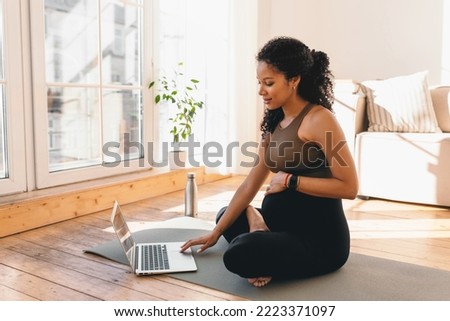 African american pregnant woman looking at laptop screen, searching for online workout tutorial or prenatal yoga sitting on floor on mat in sports wear, wearing smart watch to control heart rate Royalty-Free Stock Photo #2223371097