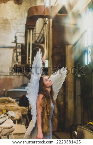 A girl with angel wings stands in the architect's studio with her head raised. Creative mess