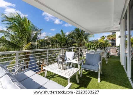 Balcony with artificial grass, overlooking the bay of Miami, white tables, chairs and armchairs, white railing, palms, blue sky and buildings in the background