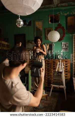 cheerful african american woman laughing and posing with tea cup near blurred man with vintage camera in art studio