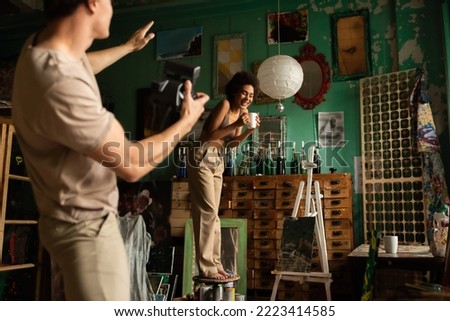 blurred man with vintage camera pointing with hand near cheerful african american woman standing on stool with cup of tea