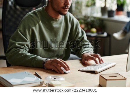 Young man in casualwear clicking mouse and pressing key of computer keyboard while working over new software by workplace Royalty-Free Stock Photo #2223418001