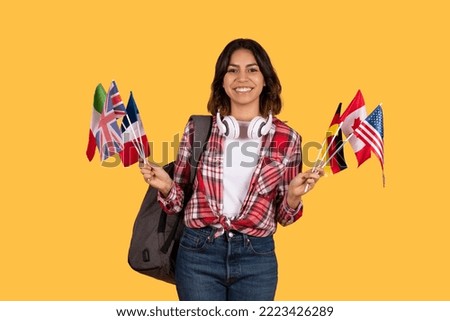 Happy pretty middle eastern young woman student holding diverse flags of various countries, carrying backpack and wireless headphones, yellow studio background, education abroad concept