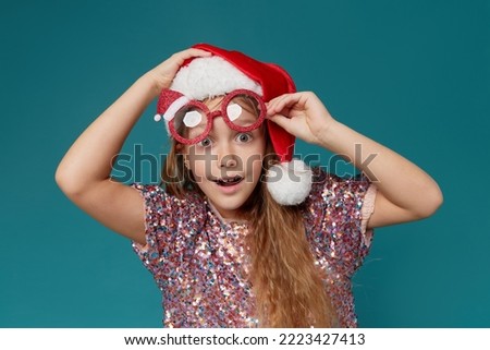 Cute little girl in a smart shiny dress in a Santa Claus hat and party glasses on a turquoise background. The child smiles in surprise with his mouth wide open. wow funny face