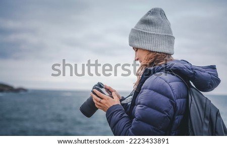 A young woman in a scarf jacket and a hat, a traveler professional photographer with a camera in her hands on the shore of the winter sea