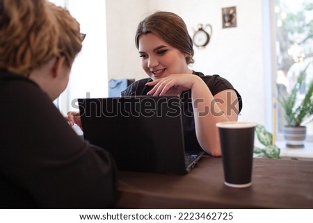 Two overweight business women sit in cafe with cup of coffee or tea and looking at the laptop, plus size female is holding a bank card to place an online order shopping