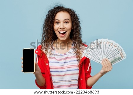 Young excited woman of African American ethnicity wear red jacket use mobile cell phone with blank screen hold fan of cash money in dollar banknotes isolated on plain pastel light blue cyan background