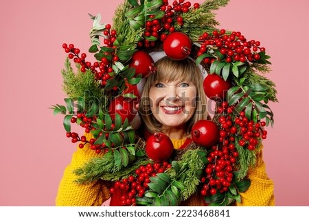 Close up merry fun smiling elderly woman 50s year old wearing yellow knitted sweater Santa hat posing hold look through wreath isolated on plain pink background Happy New Year Christmas 2023 concept