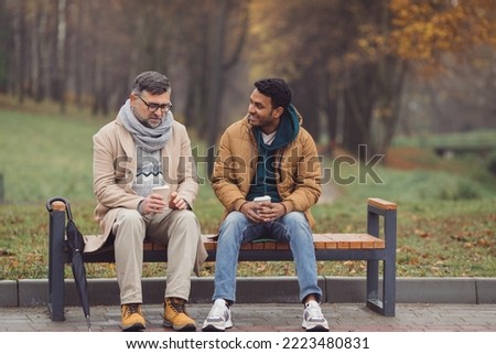 Friends, a senior and a young man sit in the park on a bench and talk in the autumn park. Royalty-Free Stock Photo #2223480831