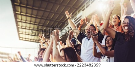 Crowd of sports fans cheering during a match in stadium. Excited people standing with their arms raised, clapping, and yelling to encourage their team to win. Royalty-Free Stock Photo #2223482359
