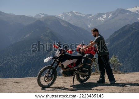 Biker man fastens dry bag on the trunk of dirt motorcycle in amazing mountainous summer landscape