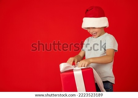 Child in a red Christmas cap curiously unties a white ribbon on a large gift box. Boy happily unties a bow from a Christmas present on a red background