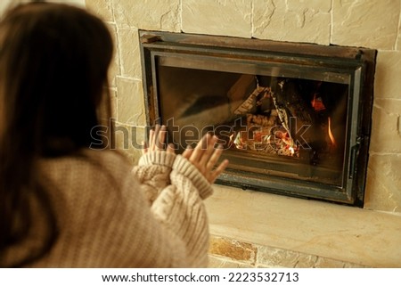Heating house in winter with wood burning stove. Woman in cozy sweater warming up hands at fireplace in rustic room. Young stylish female sitting at fireplace in farmhouse