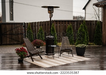 Stainless steel metal gas outdoor patio heater with wooden chairs and pot with flowers at wet terrace. Place to relax. Royalty-Free Stock Photo #2223535387