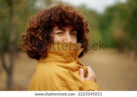 Outdoor autumn portrait of young attractive fashionable happy smiling curly girl winking covering face with orange jacket looking straight in camera. European autumn fall park photo shoot
