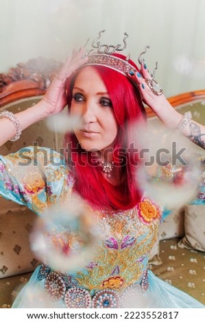 Photo of a girl in an elegant blue New Year's dress with red hair, bright makeup and a crown on her head. Beautiful apartments. Festive mood. New year's night.