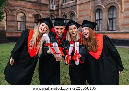 Successful graduates in academic gowns hold their diplomas, look at the camera and smile for an outdoor photo.